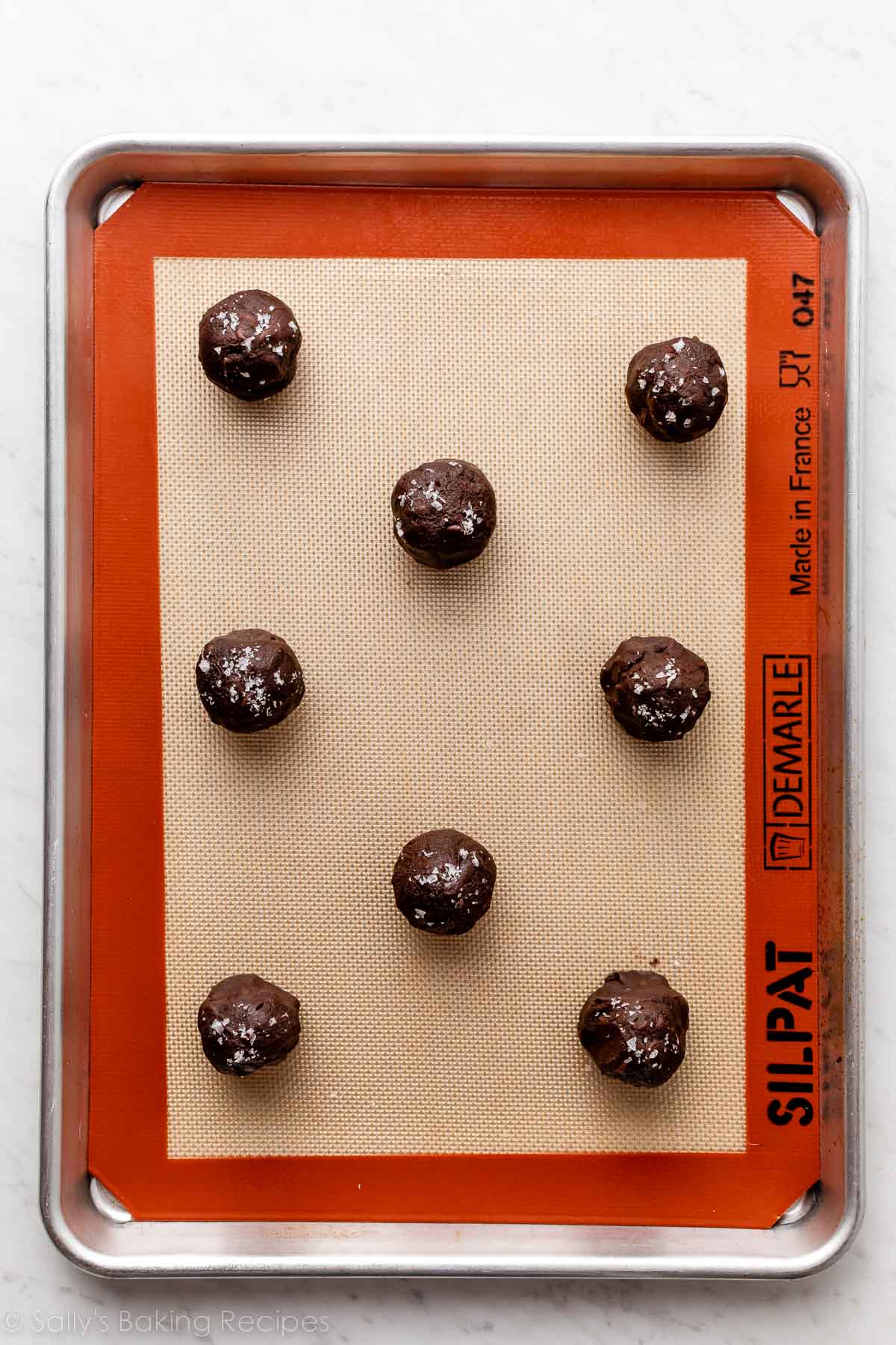 chocolate cookie dough balls with sea salt on top on silicone baking mat-lined baking sheet.