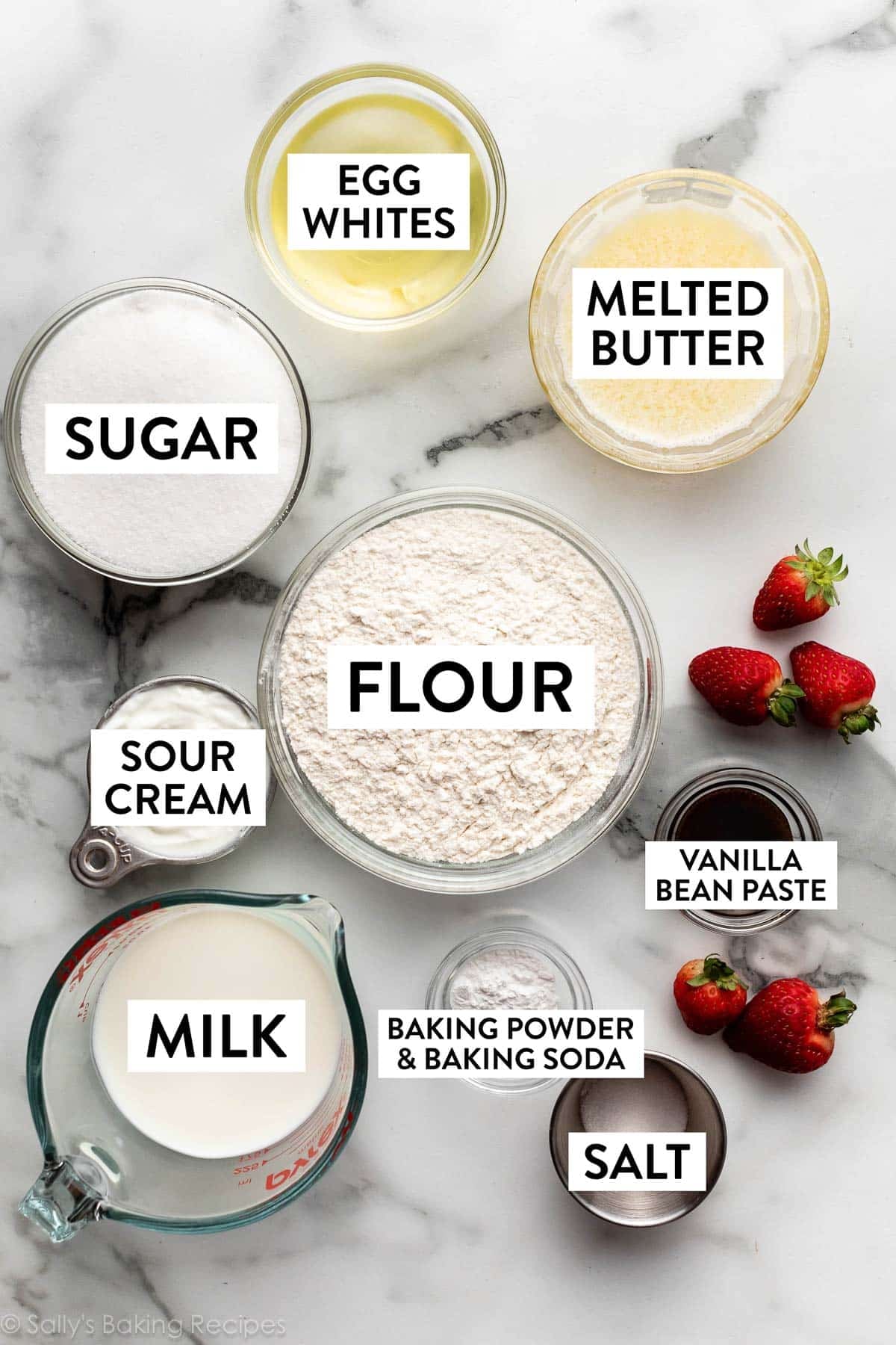 ingredients on marble counter including flour, melted butter, egg whites, strawberries, vanilla bean paste, and milk.