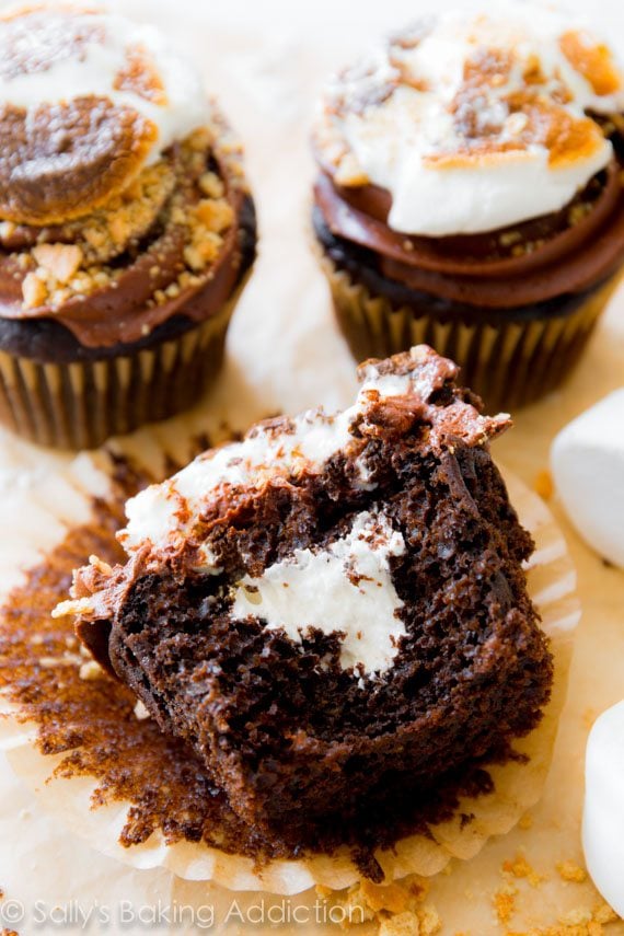 chocolate cupcakes topped with chocolate frosting, crushed graham crackers, and a toasted marshmallow with one cut in half showing the marshmallow filling
