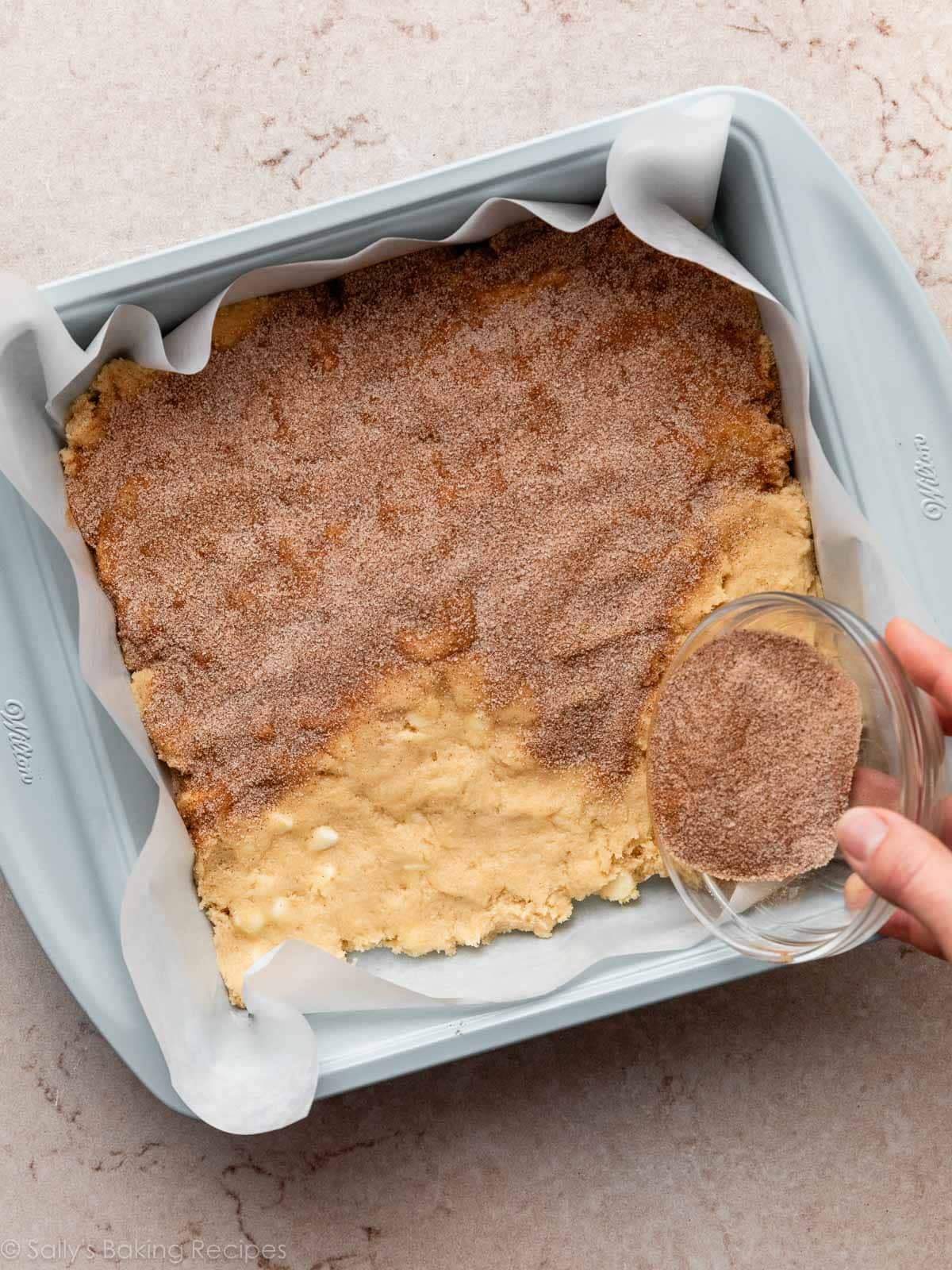 hand pouring cinnamon sugar mixture over dough in blue square pan.