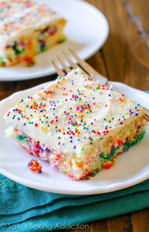 slice of funfetti sheet cake topped with vanilla frosting and sprinkles on a white plate with a fork