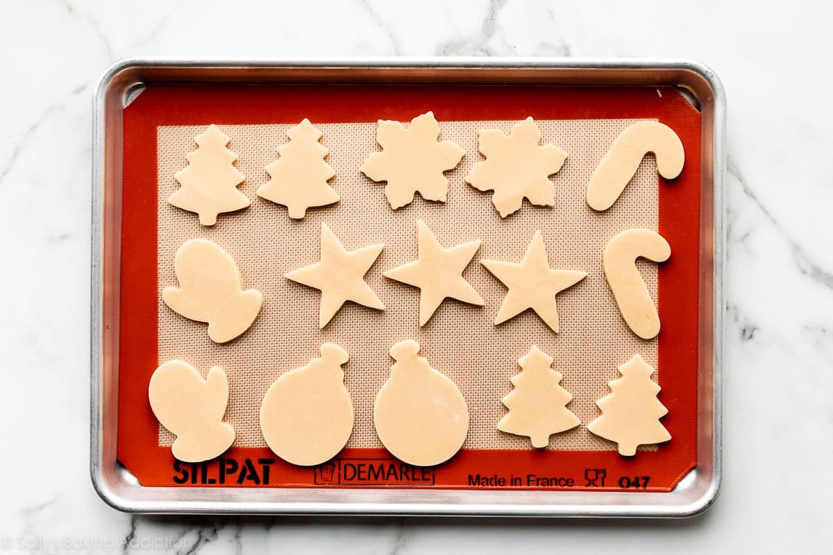 sugar cookies in various shapes on silicone mat-lined baking sheet including stars, candy canes, and snowflakes.
