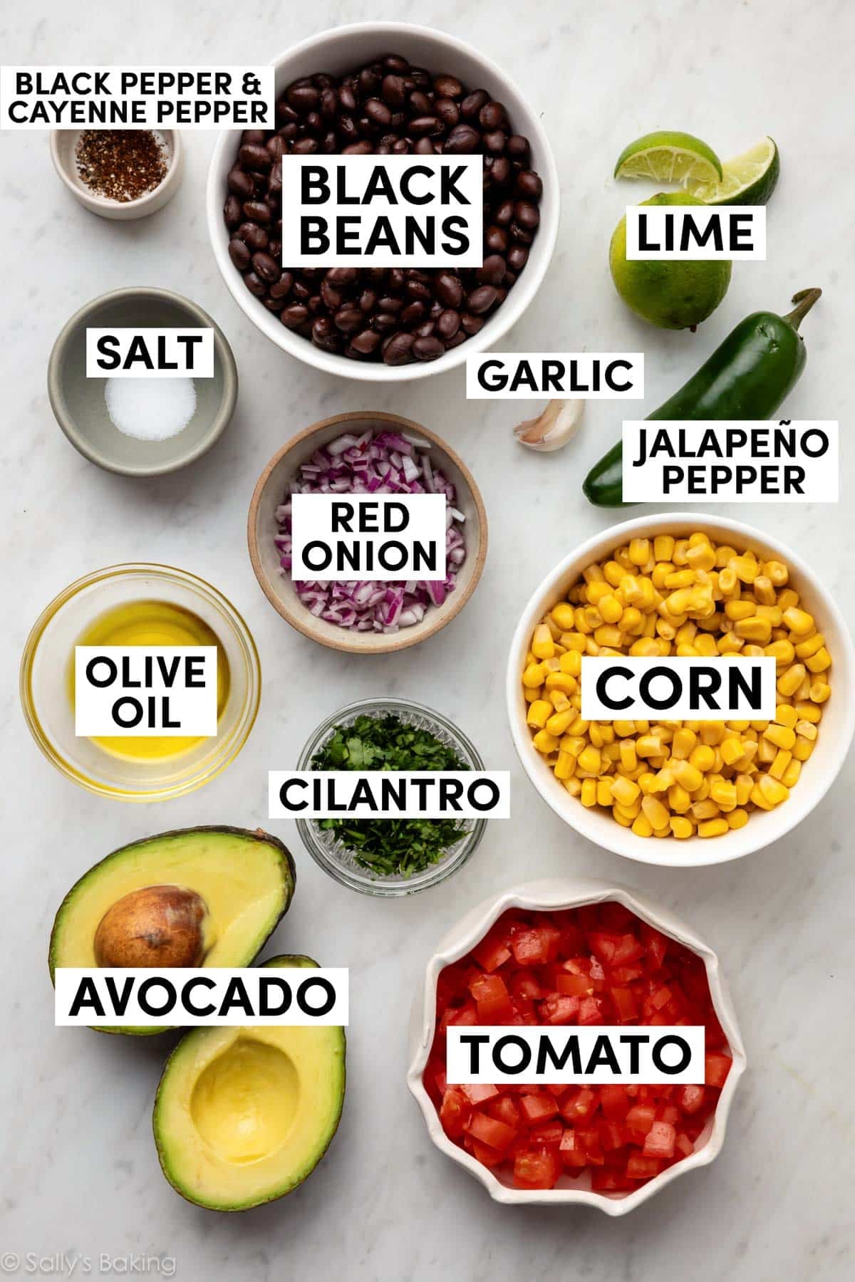 olive oil, tomatoes, red onion, lime, avocado, and other ingredients on marble backdrop.