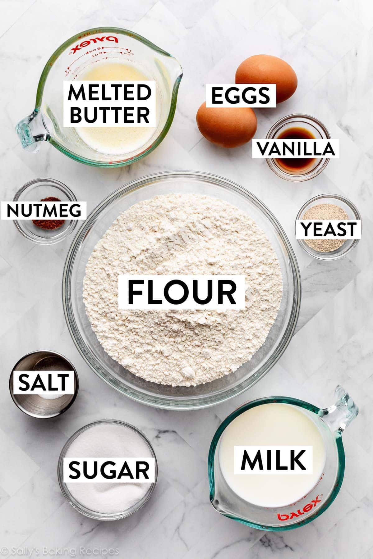 ingredients on counter including bowl of flour, sugar, salt, yeast, vanilla, nutmeg, plus 2 eggs, melted butter and milk.