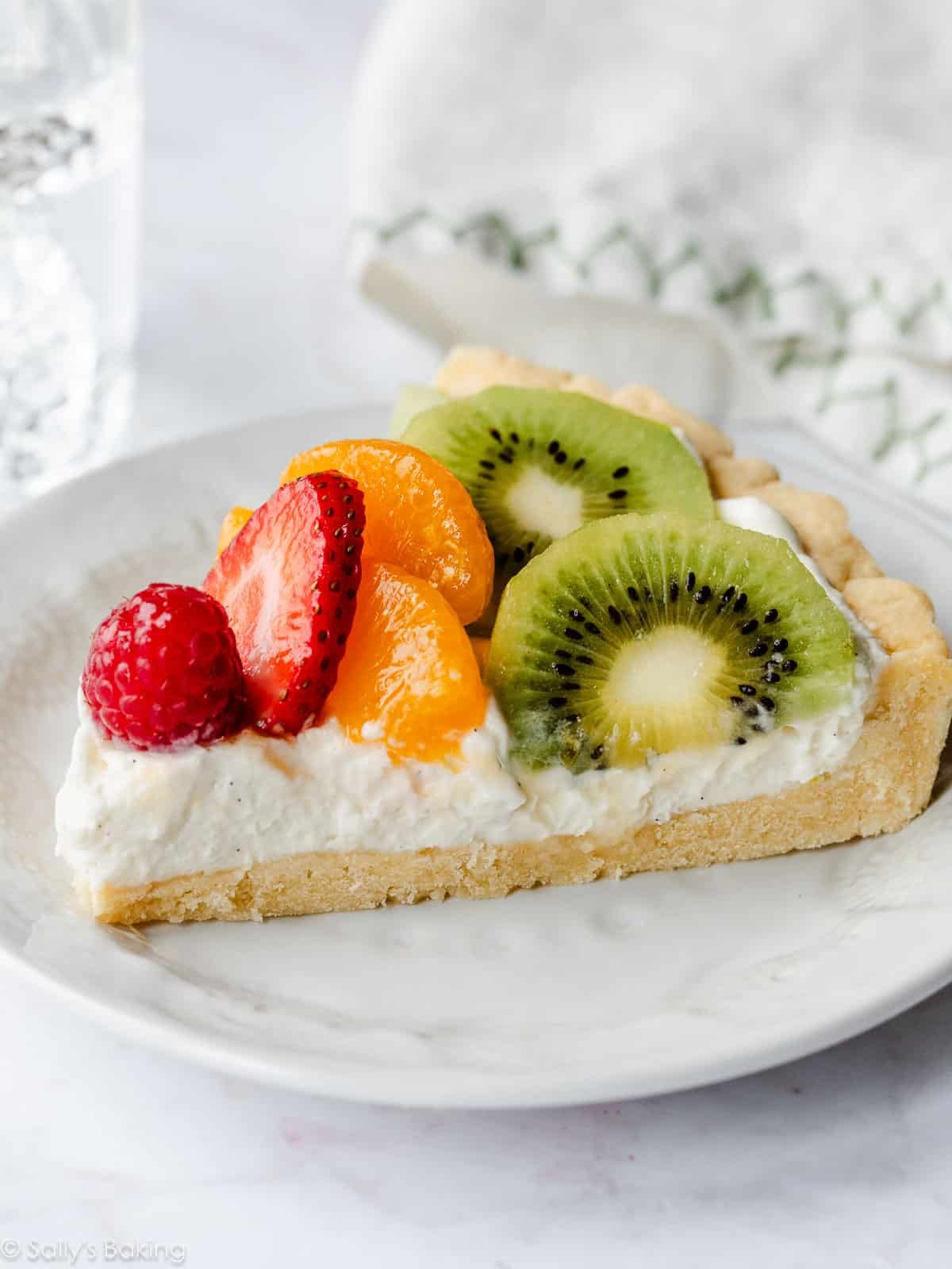 slice of fresh fruit tart with kiwi, mandarin oranges, strawberry, and raspberry on white plate.