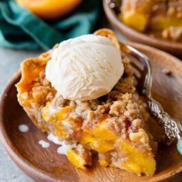 slice of peach crumble pie with a scoop of ice cream on top on a wood plate with a fork