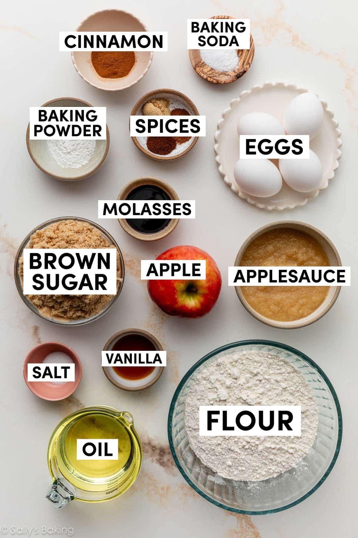 ingredients on surface including flour, applesauce, eggs, brown sugar, apple, oil, and cinnamon.