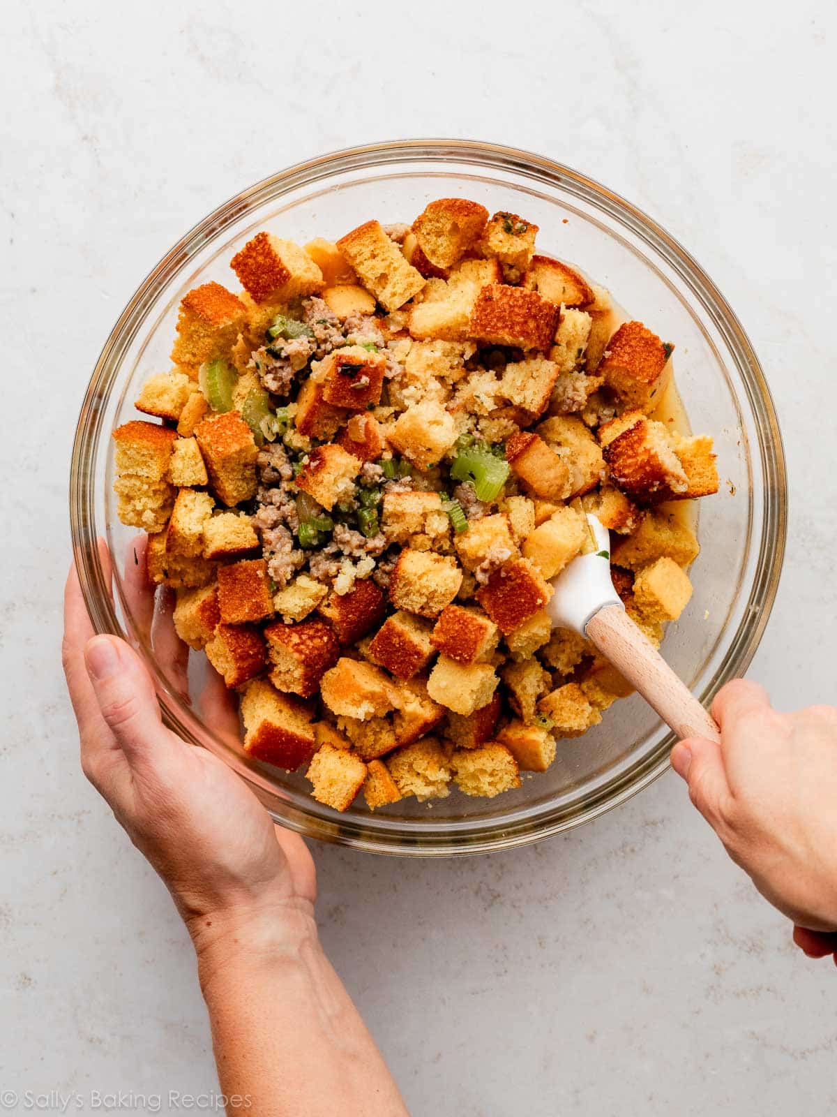 hands folding bread cubes, sausage, and herbs together in glass bowl.