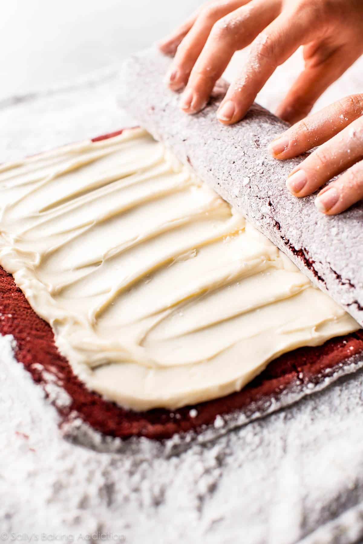 hands rolling up red velvet sponge cake with cream cheese frosting