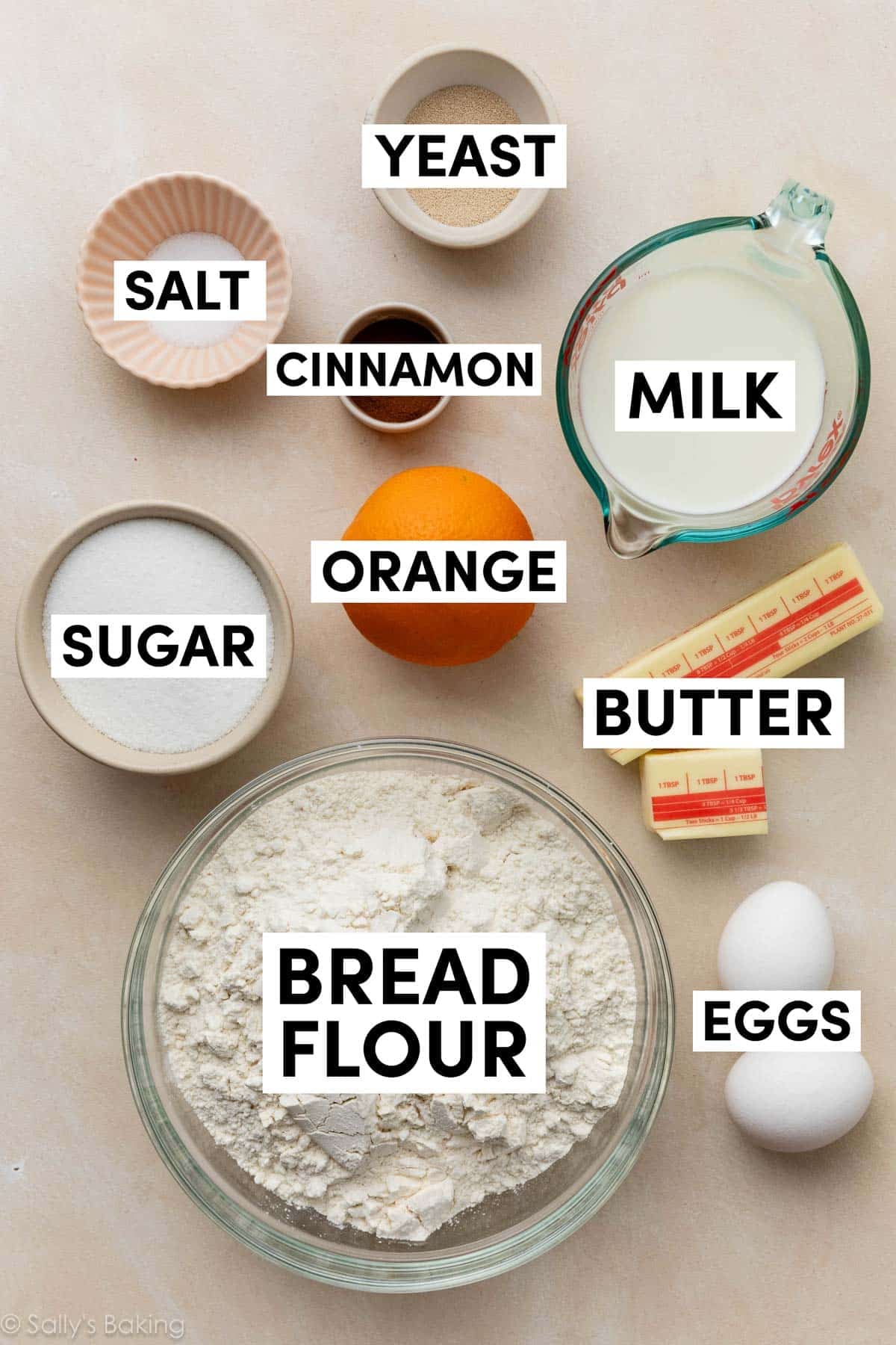 ingredients on beige surface including bread flour, butter, milk, eggs, cinnamon, sugar, and yeast.