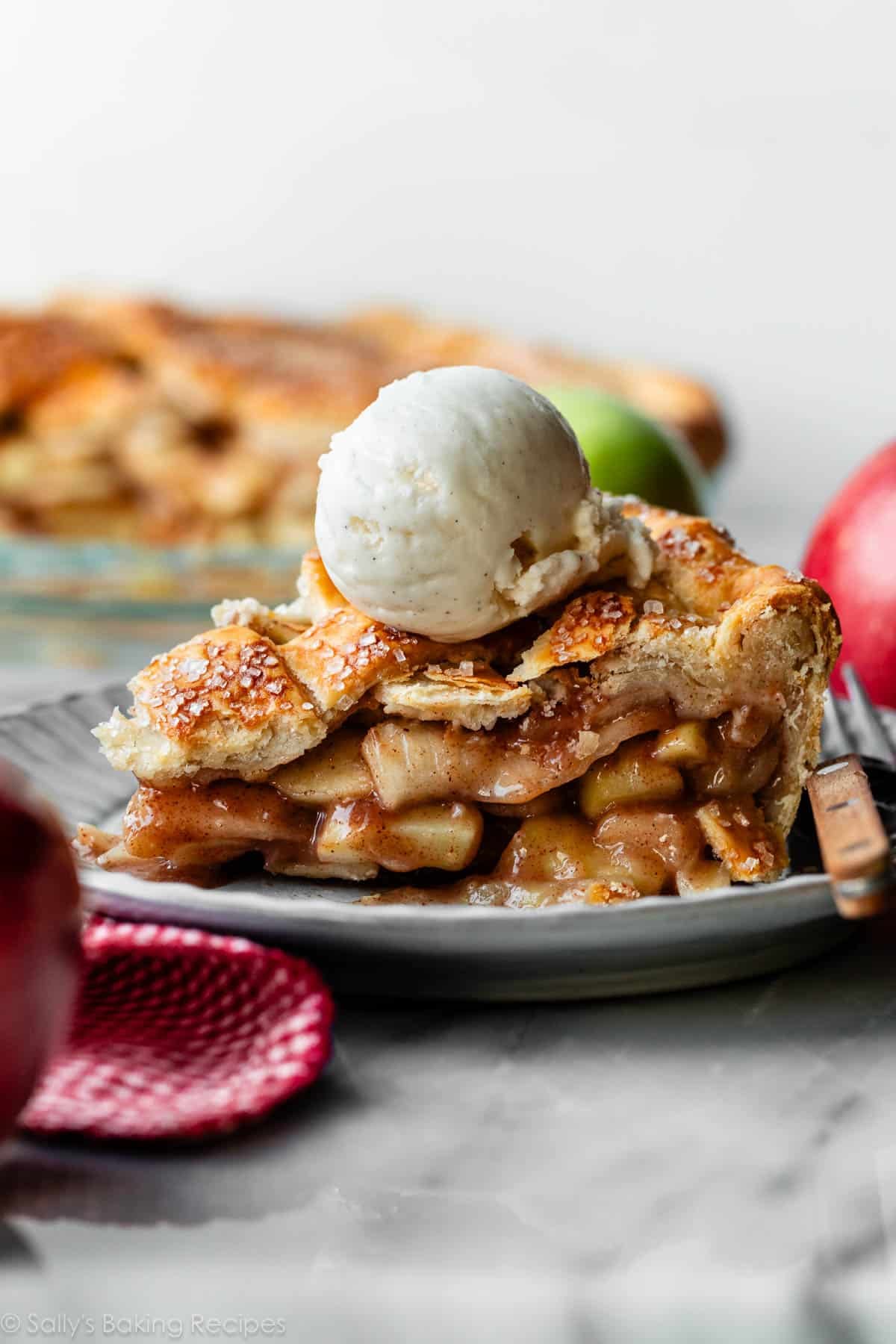 apple pie slice on plate with a scoop of vanilla ice cream on top and apples in the background.