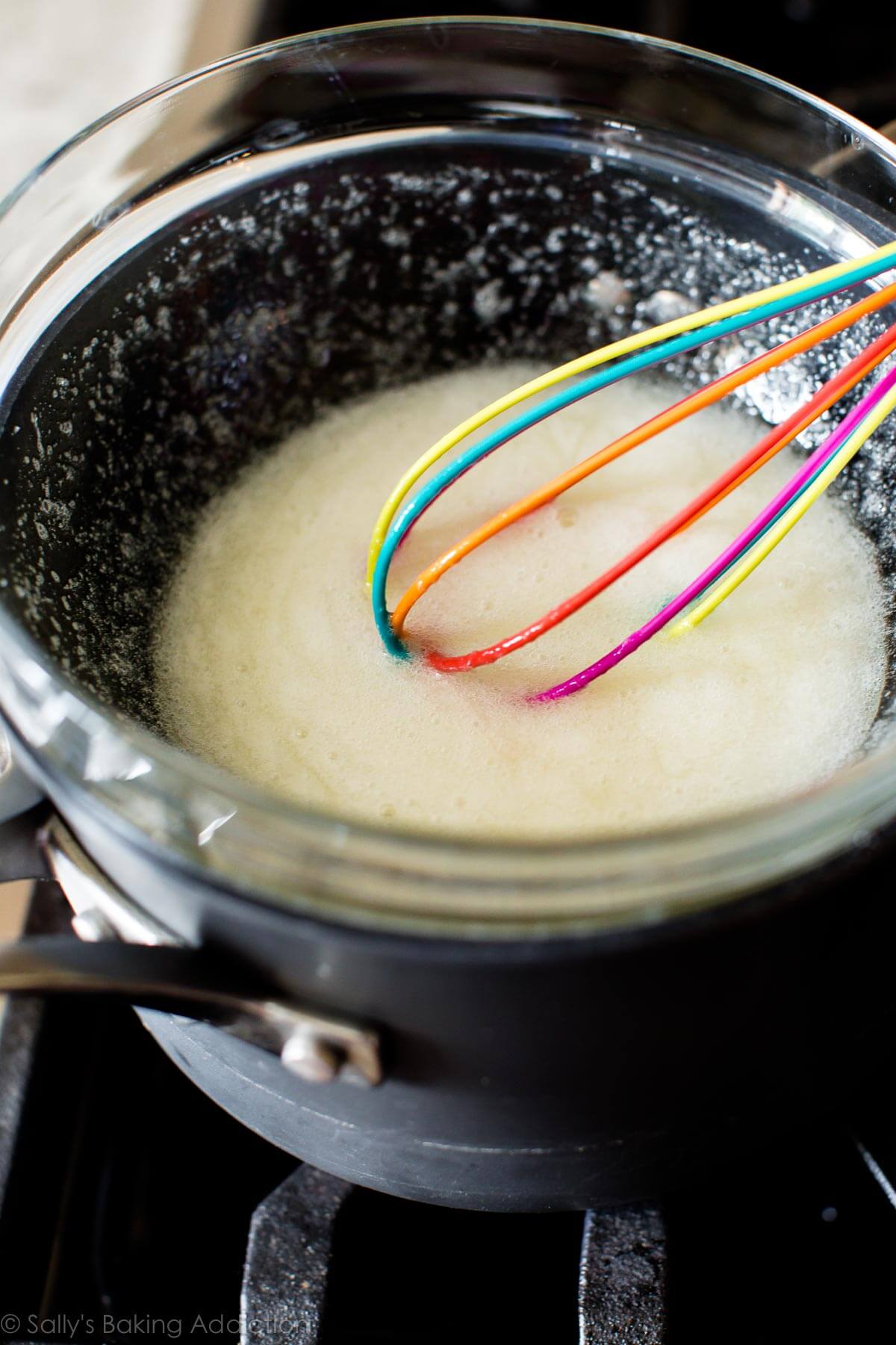 egg whites, sugar, and cream of tartar whisked together in a glass bowl on top of a pot of simmering water on the stovetop