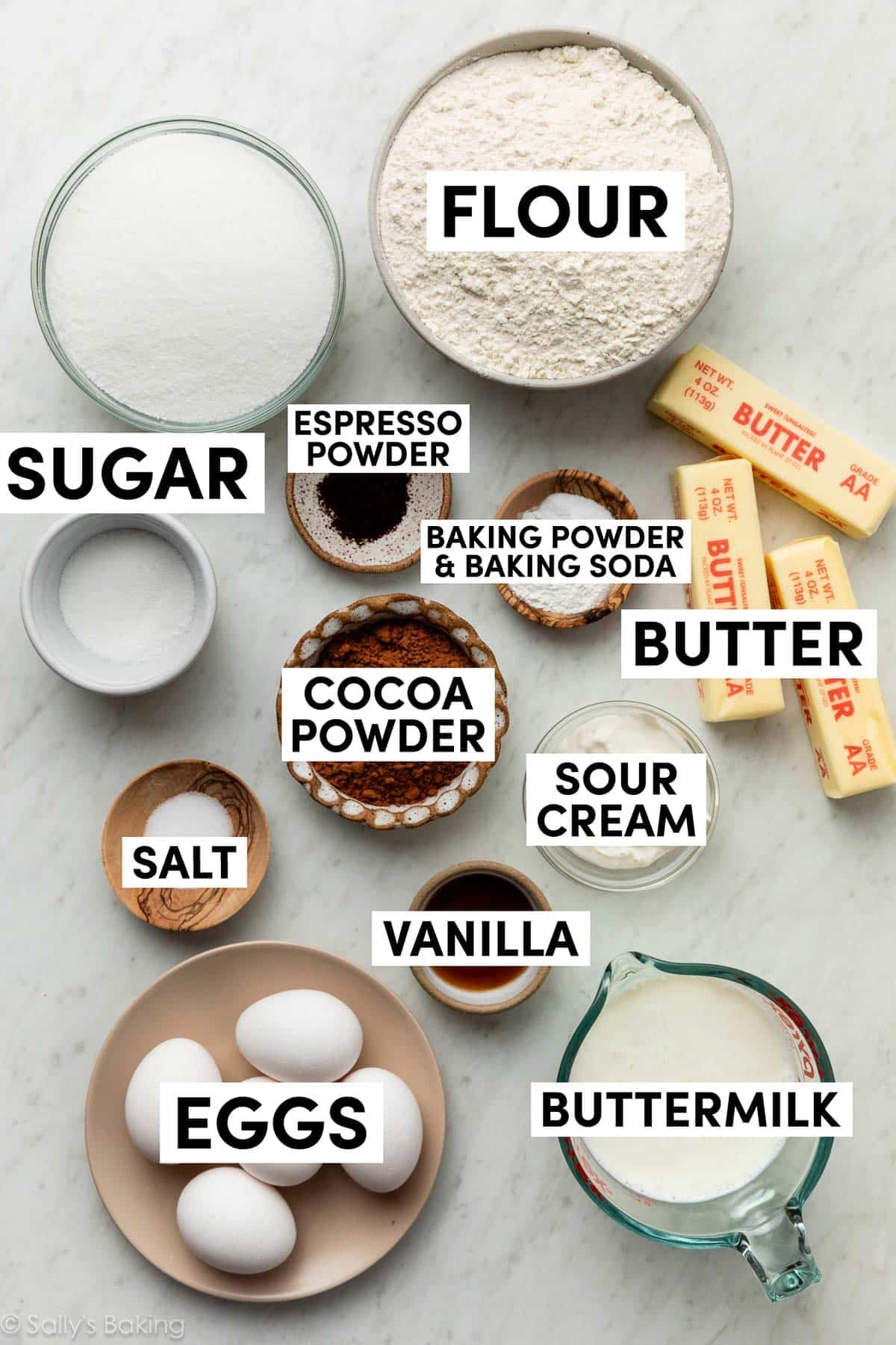 ingredients on marble surface including eggs, butter, flour, buttermilk, cocoa powder, sour cream, vanilla, and sugar.