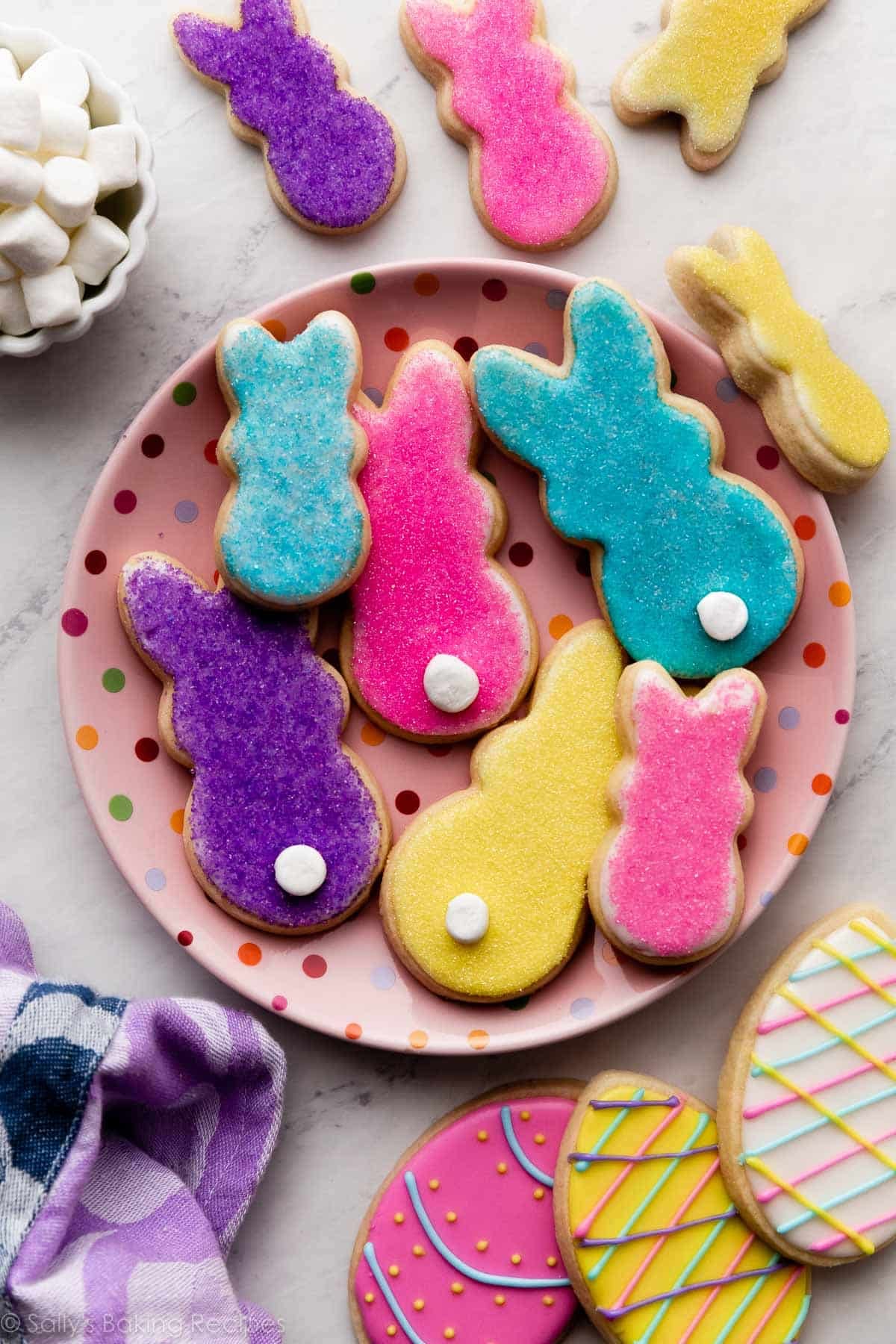 PEEPS marshmallow-inspired bunny sugar cookies with marshmallow tails on pink polka dot plate with Easter egg decorated sugar cookies in bottom corner.