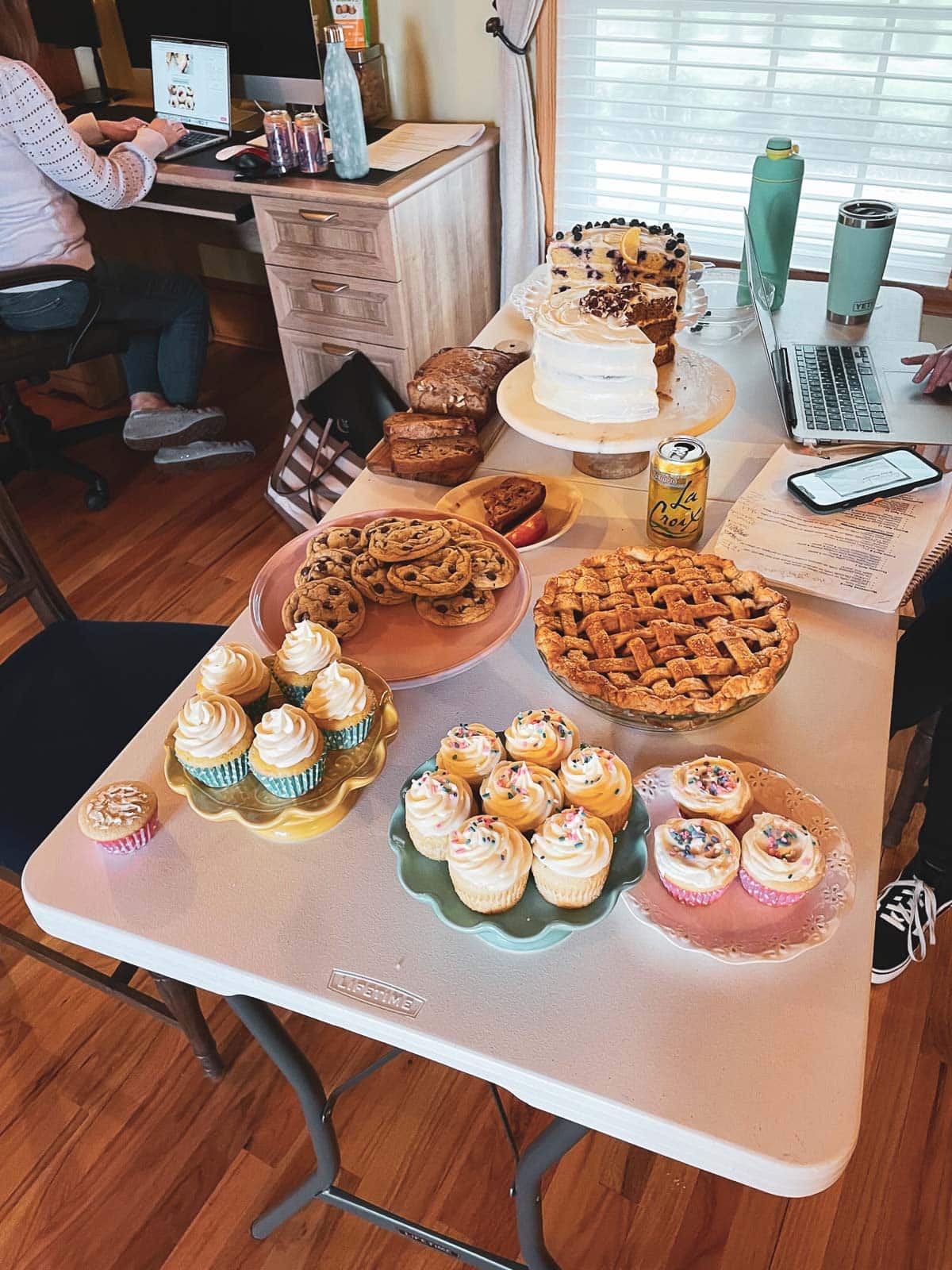 many different baked goods on table including apple pie, cupcakes, chocolate chip cookies, and carrot cake.