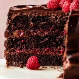chocolate raspberry cake cut open with raspberries and mint on top sitting on a marble cake stand with pink backdrop behind it.