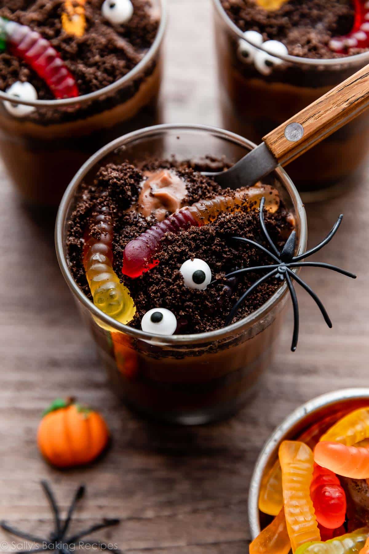 chocolate dirt pudding cups for Halloween with a pretend spider on top, gummy worms buried in crushed Oreos, and a spoon scooping out the 1st bite.