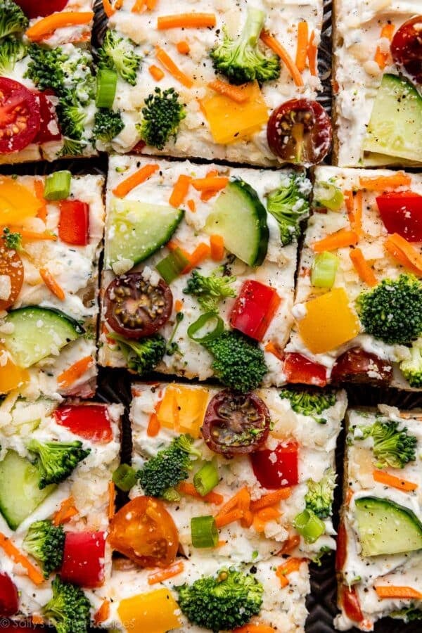 overhead photo of cream cheese veggie pizza with broccoli, red pepper, yellow pepper, cherry tomatoes, shredded carrots, cucumber, and parmesan cheese on top.