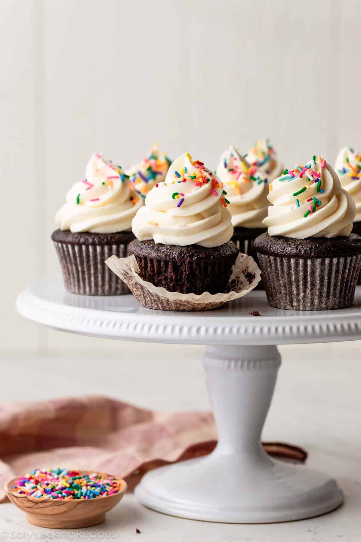 white cake stand with chocolate cupcakes with vanilla buttercream on top and decorated with rainbow sprinkles.