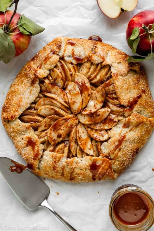 apple galette with caramel sauce on white parchment paper with apples and pie server around it.