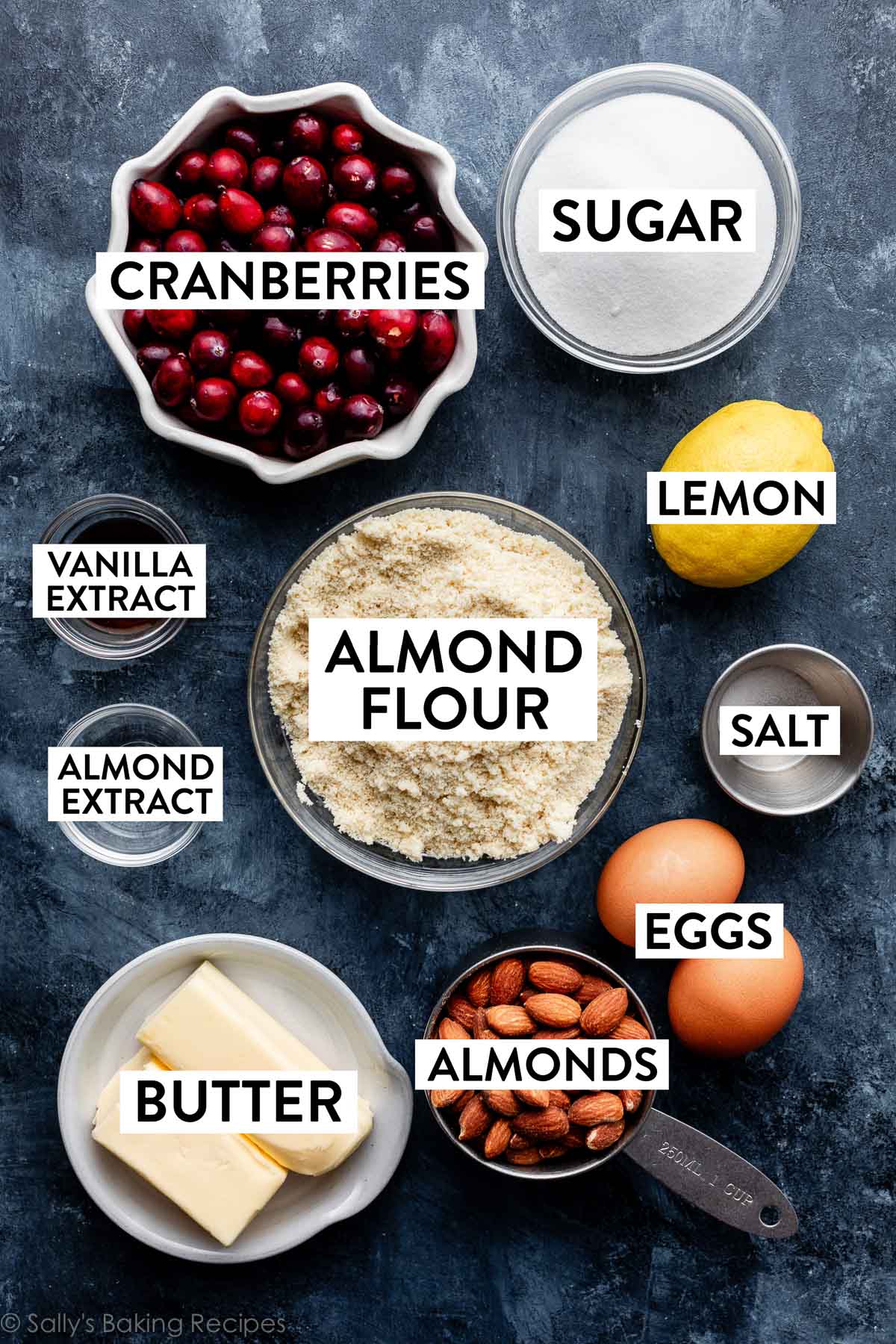 bowls of measured ingredients on blue surface including sugar, fresh cranberries, almonds, almond flour, butter, vanilla extract, almond extract, and salt.