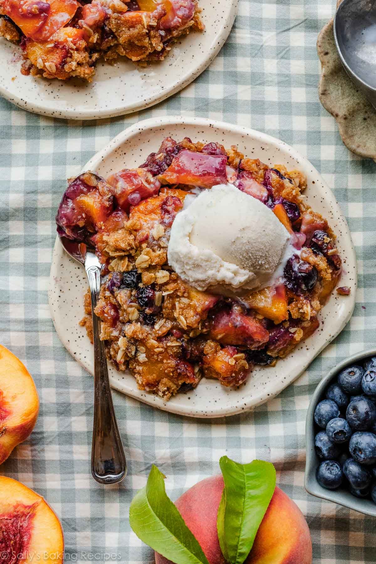 blueberry peach crisp with brown butter oat topping and vanilla ice cream on plates on blue gingham linen.