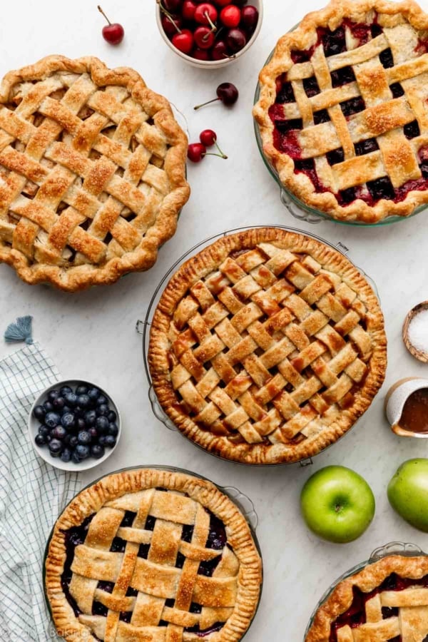 pies with lattice pie crust on marble surface including blueberry pie, 2 apple pies, and cherry pie.