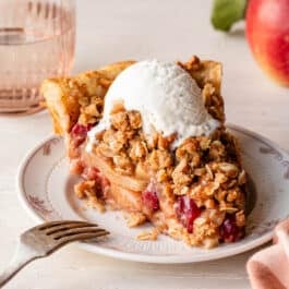 slice of apple cranberry crumb pie on white plate with vanilla ice cream on top and pink glass in the background.