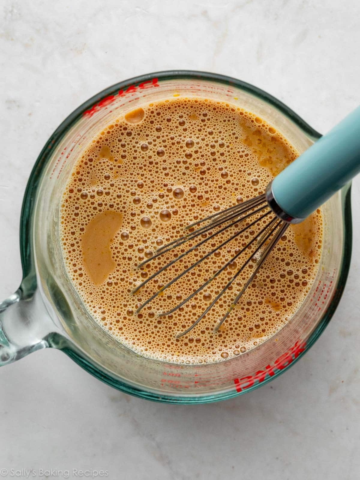 brown liquid mixture in large liquid measuring cup with blue-handled whisk.