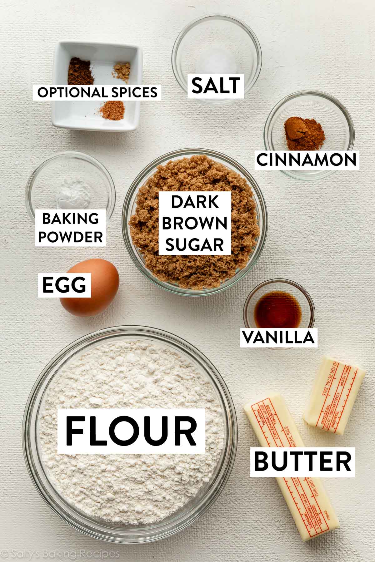 ingredients on white surface including butter, flour, vanilla, egg, brown sugar, spices, and salt.