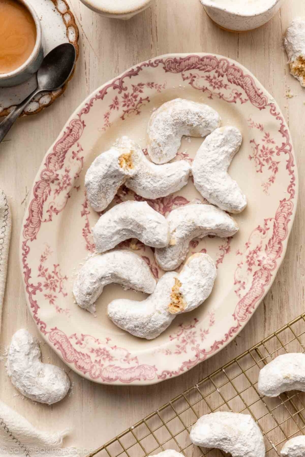 almond crescent cookies on pink floral plate with white linen and coffee next to it.