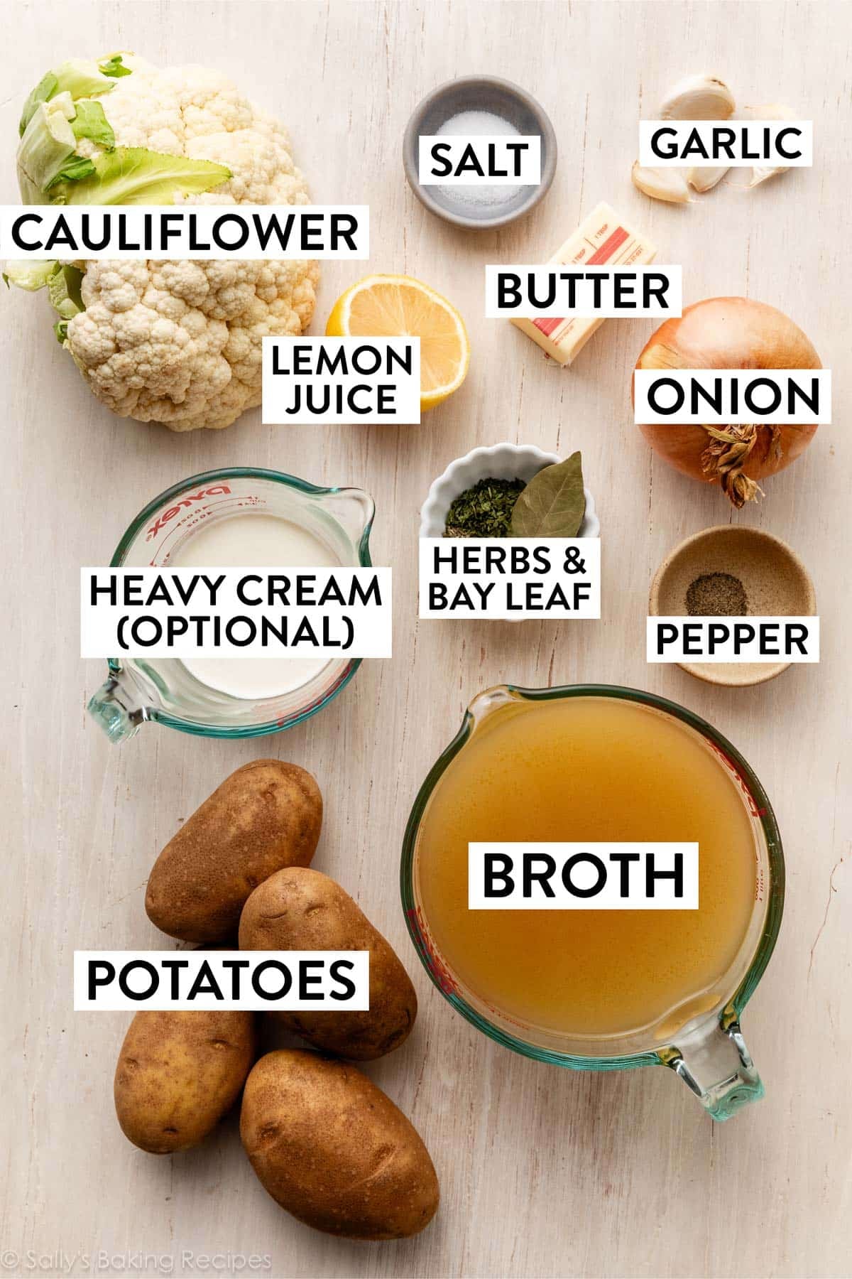 ingredients on white wooden surface including broth, herbs, bay leaf, pepper, onion, salt, garlic, lemon, and potatoes.
